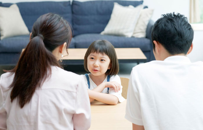 little Asian girl facing two adults across a table with adults' backs to the camera