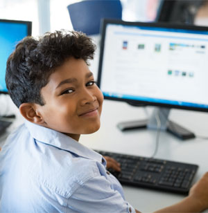 boy sitting in front of a computer monitor and looking over his shoulder to smile at camera